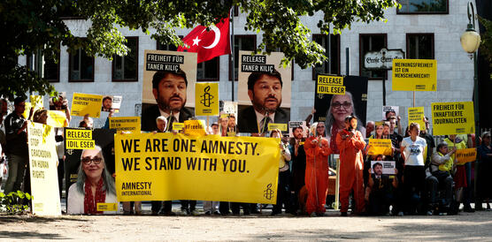 Menschen vor weißem Gebäude mit Türkei-Flagge, die gelbe Amnesty-Banner sowie Schilder mit der Forderung "Release İdil Eser and Taner Kılıç" hochhalten 