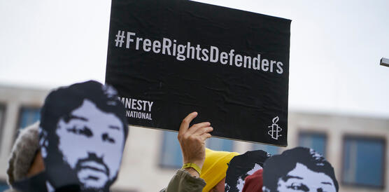 Personen halten Papp-Masken mit dem Gesicht von Taner Kılıç und ein Schild mit dem Schriftzug "#FreeRightsDefenders"
