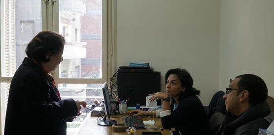 Eine ältere Dame spricht im Stehen mit einer jüngeren Frau und einem jüngeren Mann, die an einem Schreibtisch mit Comuter in einem engen Büro sitzen
