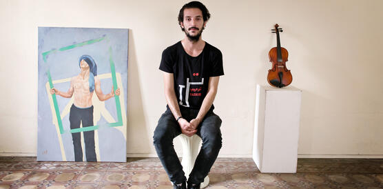 Ein junger Mann sitzt auf einem Hocker, hinter ihm links ein Gemälde und rechts eine Geige
