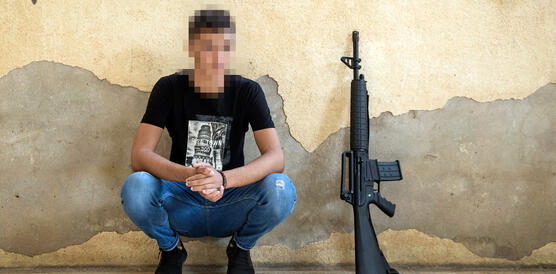 Ein junger Mann hockt vor einer Wand, neben ihm ein Maschinengewehr