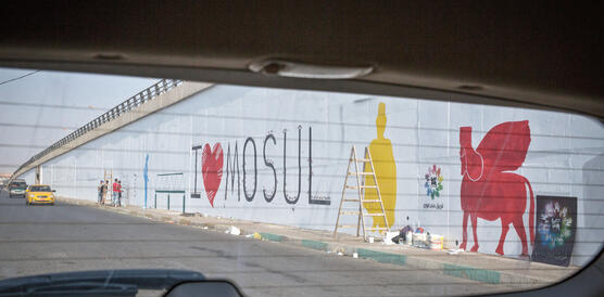 Aus dem Auto sieht man eine Straße in Mossul, wo vier junge Männer an eine Mauer malen