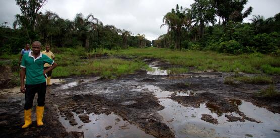 Drei Männer in Gummistiefeln stehen in einem schlammigen Gebiet im Nigerdelta, das von der Ölverschmutzung betroffen ist