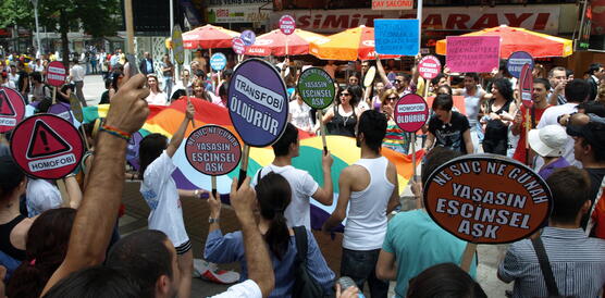 Menschen auf der Straße, halten Schilder, in der Mitte eine große Regenbogenflagge