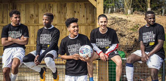 Vier Jungs sitzen in Fußballtrikots auf einem Zaun, vorne mittig steht einer mit einem Ball in der Hand und lacht
