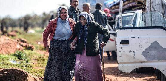 Zwei syrische Frauen steigen aus einem Auto, eine von ihnen stützt die andere. Im Hintergrund warten weitere Personen mit Fahrzeugen.