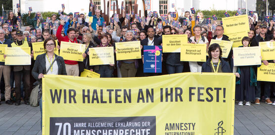 Eine Gruppe von Menschen hält gelbe Schilder hoch, auf denen jeweils ein Menschenrecht geschrieben steht. Im Vordergrund halten zwei Frauen ein großes Banner, auf dem "Wir halten an ihr fest! 70 Jahre Allgemeine Erklärung der Menschenrechte"