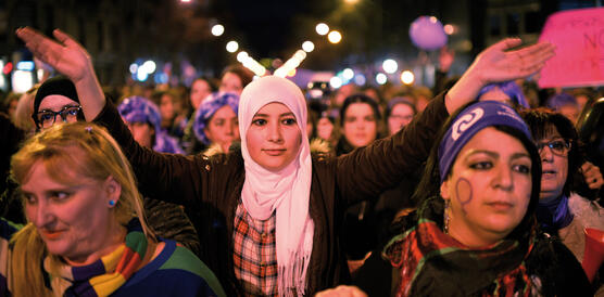 Viele Frauen laufen bei einer Demo, eine Frau mit weißem Kopftuch in der Mitte streckt die Arme zum Klatschen in die Luft