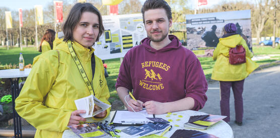 Eine junge Frau in gelber Amnesty-Jacke und ein junger Mann stehen an einem Stehtisch mit Flyern und lächeln in die Kamera