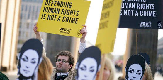Eine Gruppe von Personen steht vor dem Brandenburger Tor und trägt Pappmasken mit dem Gesicht von Eren Keskin und hält Schilder und Banner in den Händen. Im Fokus ist ein Schild mit der Aufschrift „Defending human Rights is not a crime“