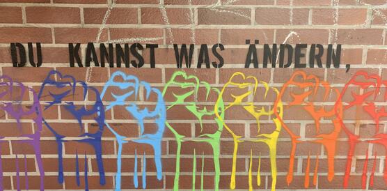 Graffito mit vielen regenbogenfarbenen Fäusten und der Aufschrift "Du kannst was ändern, als tu es!" auf einer roten Backsteinmauer