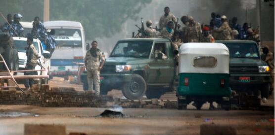 Mehrere bewaffnete Soldaten blicken auf ihren Autos stehend auf eine niedergebrannte Fläche auf der Straße. 