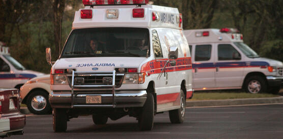 Zwei Rettungskräfte sitzen in einem Krankenwagen, der auf einem Parkplatz steht, im Hintergrund stehen weitere Krankenwagen und ein geparktes Auto