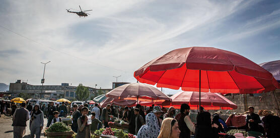 Ein Marktplatz mit Menschen, im Vordergrund ein Sonnenschirm, am Himmel ein Hubschrauber