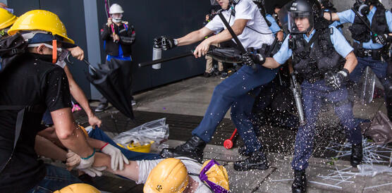 Zwei Demonstrierende helfen einem am Boden liegenden Demonstraten auf, während eine Gruppe von Sicherheitskräften in Schutzausrüstung auf sie zustürmt und der Polizist den am Boden liegenden Mann tritt
