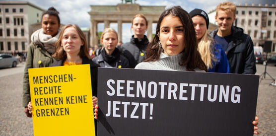 Vor dem Brandenburger Tor stehen zwei Männer und fünf Frauen. Zwei Frauen im Vordergrund halten jeweils ein Plakat. Auf dem linken steht: "Menschenrechte kennen keine Grenzen" und auf dem rechten steht: "Seenotrettung jetzt!"