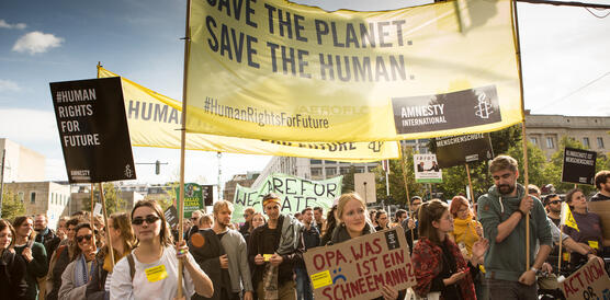 Eine protestierende Menschenmasse mit Bannern und Schildern, die friedlich demonstrieren. Auf dem gelben Amnesty-Banner steht "Save the planet, save the human".