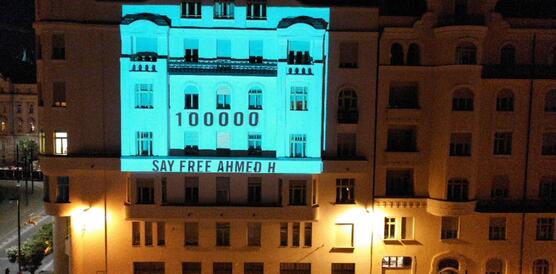 Auf einem Gebäude, welches wegen der Tageszeit beleuchtet ist, wird ein Bild projiziert, auf dem steht: "100.000 Menschen fordern: Lasst Ashmed H. frei"