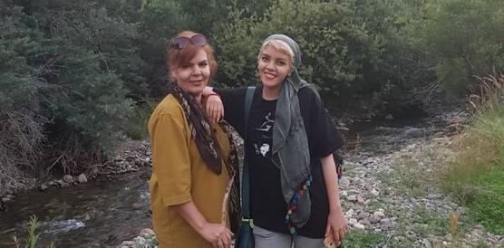 Die beiden Frauen stehen an einem Fluss, im Hintergrund ist Gebirge zu sehen. Sie stehen dicht beisammen und lächeln in die Kamera.