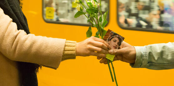 Eine Hand übergibt eine Blume mit einem Kärtchen in die Hand einer anderen Person. Im Hintergrund fährt die U-Bahn ein.