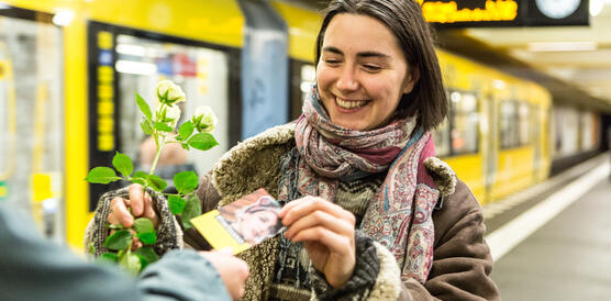 Eine junge Frau bekommt in einer Berliner U-Bahnstation eine Blume und ein Kärtchen mit einem Frauenporträt überreicht. Sie lacht dabei. Im Hintergrund fährt eine gelbe U-Bahn vorbei.