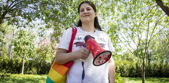 Frau mit Megaphon und Tasche in Regenbogenfarben steht an einem sonnigen Tag in einem Park