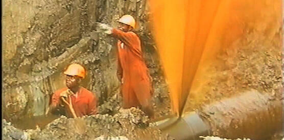 Zwei Arbeiter in orangenen Uniformen und Helmen neben einer Pipeline, aus der ein Ölstrahl spritzt