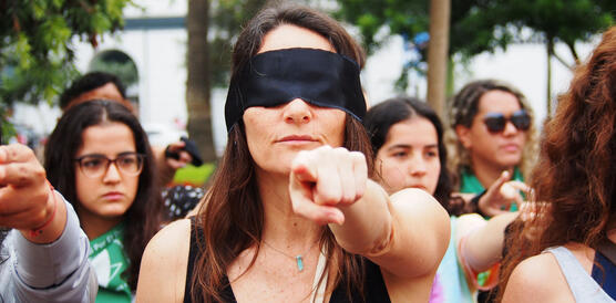 Mehrere Frauen und Mädchen demonstrieren, im Vordergrund eine Frau mit schwarzer Augenbinde, sie zeigt direkt in die Fotokamera
