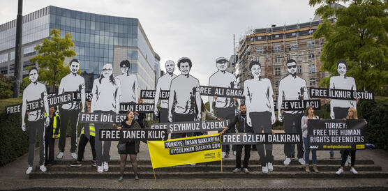 Mehrere Personen demonstrieren, stehen auf einer Treppe mit mehreren knapp drei Meter großen Darstellungen von inhaftieren Menschenrechtsverteidiger_innen