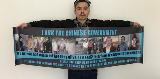 Ein Mann hält ein großes Plakat mit Fotos von mehreren Personen, auf dem "I ask the Chinese government: My parents and relatives! Are they alive or dead? In prison or in concentration camps? Please God, help me to save my family, make our sorrows and voice to be heard by the people of peace and love around the world." steht