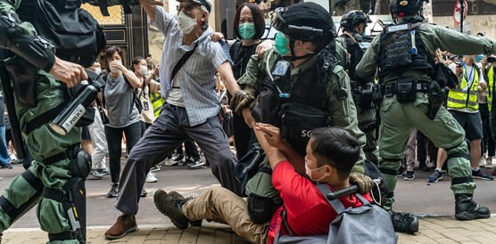 Vor dem Hintergrund einer Demonstration, drückt ein Polizist einen Demonstranten zu Boden. Daneben greift ein älterer Mann einen Polizisten an.
