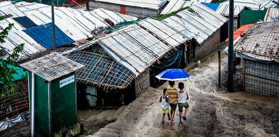 Drei Kinder, von hinten fotografiert, laufen unter einem großen Regenschirm einen Weg zwischen Hütten aus Metallgittern und Plastikfolien entlang