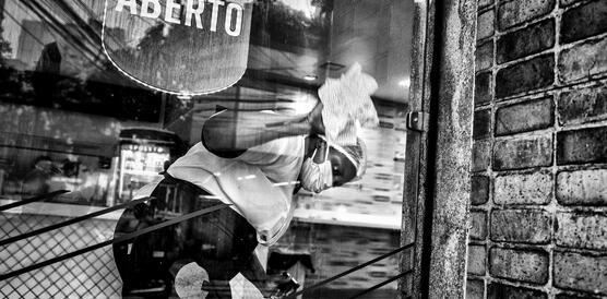 Eine junge Frau mit Mundschutz und Kopfhaube, es ist die Brasilianerin Lívia Nogueira, steht in einem Laden und putzt die Schaufensterscheibe von innen. 