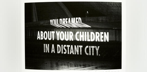 Schwarzweißfoto eines Kunstwerks der Künstlerin Jenny Holzer, die einen weißen Schriftzug (You dreamed about your children in a distant city) auf eine dunkle Fläche projiziert.