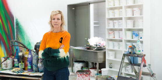 Eine mittelalte Frau mit blonden Haaren, es ist die Künstlerin Katharina Grosse, trägt einen bunten Pulli und steht mit ernstem Blick und verschränkten Armen in ihrem Atelier vor Farbtöpfen und einer Leinwand.