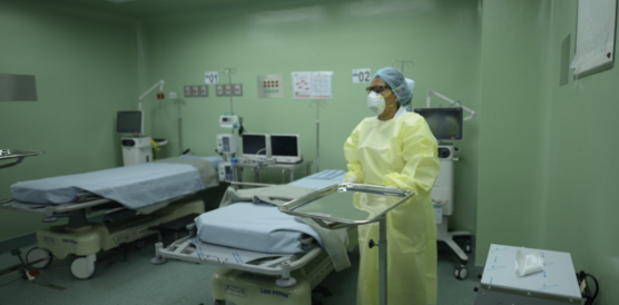 Ein Zimmer in einem Krankenhaus mit grünlichen Wänden und zwei Betten. Eine Frau mit gelbem Schutzanzug und Maske rechts im Bild verschiebt einen Tisch für medizinisches Besteck.