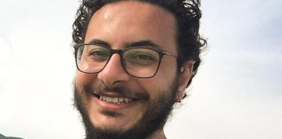Porträt eines Mannes mit dunklen Locken und Brille, der in die Kamera lächelt.