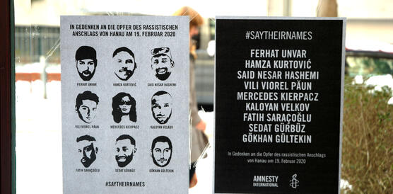 Foto von zwei Plakaten, die in einem Fenster hängen. Im Hintergrund sieht man eine beschneite Straße. Auf einem Plakat sind die Gesichter der neun Menschen abgebildet, die bei dem rassistische Attentat in Hanau am 19.02.2020 ermordet wurden. Auf dem anderen Plakat sind ihre Namen aufgelistet.