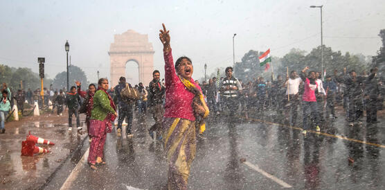 Eine Frau läuft vorm India Gate in Neu-Delhi die Straße entlang. Sie hat ihre rechte Hand erhoben und ruft etwas. Dabei treffen sie Wassertropfen, potenziell von einem Wasserwerfer. Hinter ihr befinden sich weitere Protestierende.