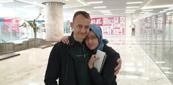 Hüseyin Galip Küçüközyiğit und seine rechts neben ihm stehende Tochter umarmen sich und blicken in die Kamera. Sie stehen allein in einem breiten, hell erleuchteten Flur in einem Einkaufszentrum oder einem Flughafen.