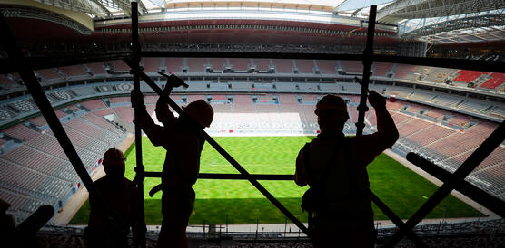 Das Bild zeigt das Innere eines Fußballstadion, im Vordergrund die Silhouetten mehrere Bauarbeiter, die ein Baugerüst abbauen
