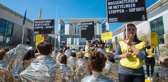 Das bild zeigt Demonstrierende mit Rettungsfolien, die Schilder halten mit Aufschriften wie "Massensterben im Mittelmeer stoppen"