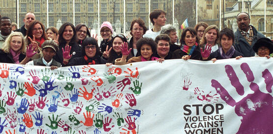Das Bild zeigt Demonstrierende und ein Plakat mit vielen bunten Händen und der Aufschrift "Stopp gegen Gewalt an Frauen"