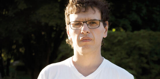 Ein junger Mann mit Brille und V-Neck-T-Shirt blickt ernst in die Kamera.