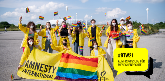 Eine Menschengruppe in gelben Jacken wedeln mit Regenbogenflaggen, sie haben vor sich ein gelbes Transparent auf dem ebenfalls die Regenbogenflagge ist und auf dem "Amnesty International" steht. Im Hintergrund die Allianz-Arena in München. Rechts unten in der Ecke eine gelbe Sprechblase, darin steht in schwarzer Schrift: #BTW21. Komromisslos für Menschenrechte