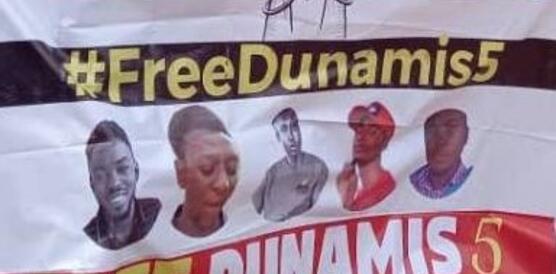 Zu sehen ist ein Banner mit der Aufschrift "Free Dunamis" und den Portraits der fünf Aktivisten, das offensichtlich von mehreren Personen getragen wird (von denen nur Hände und Füße zu sehen sind).