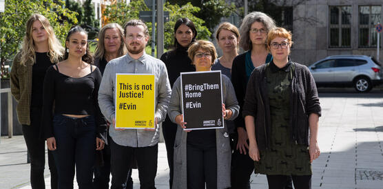 Das Foto zeigt eine Gruppe von neun Personen auf einem Bürgersteig, die ernst in die Kamera blicken. Zwei von ihnen halten Schilder hoch, auf denen steht: "This it not justice! #Evin" und "Bring them home! #Evin".