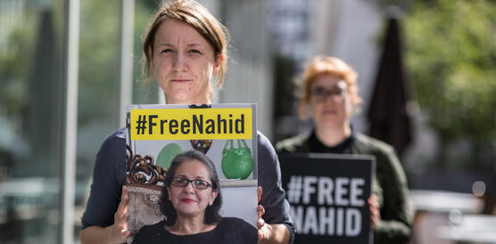 Das Foto zeigt eine Frau auf einem Bürgersteig, die in die Kamera blickt und ein Schild vor sich hält, auf dem steht: "#FreeNahid".