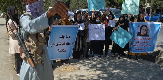 Das Bild zeigt mehrere Frauen mit Plakaten im Hintergrund, im Vordergrund ein Taliban-Kämpfer mit schwerer Bewaffnung