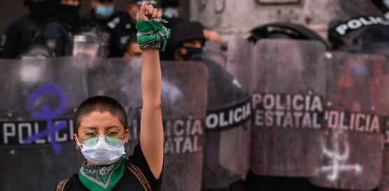 Eine junge Frau mit Maske vor dem Mund reckt eine Faust in die Höhe, dahinter Polizisten mit Schildern.